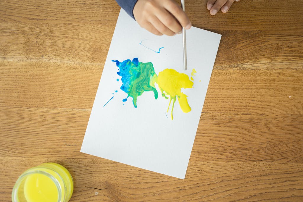 From above of crop unrecognizable kid drawing with watercolors using straw on wooden table with glass jar of water