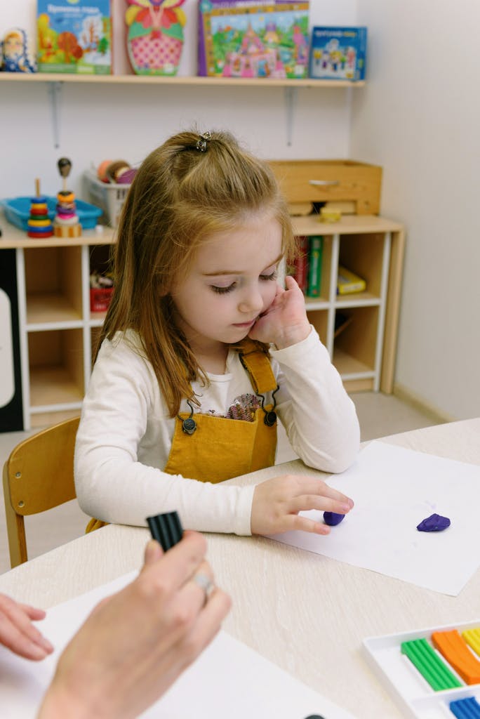 Girl in White Long Sleeve Shirt Sitting at Table and Sculpting of Clay